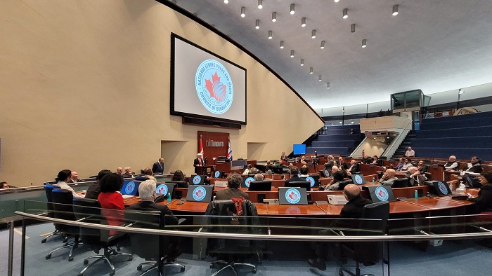 Formal council meeting in a large chamber, delegates seated at curved desks with laptops and microphones, a large screen displaying a circular emblem at the front.