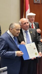 Three men in suits at a formal ceremony; the man on the left holds a blue folder with a certificate.