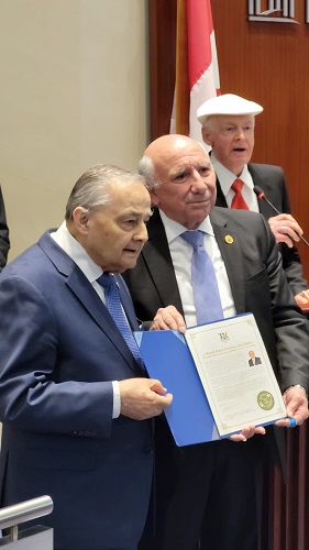 Three men in suits at a formal ceremony; the man on the left holds a blue folder with a certificate.