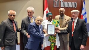 Group of six people at an award ceremony, the woman in green and yellow receives a framed certificate from two men, with a Toronto backdrop behind them.