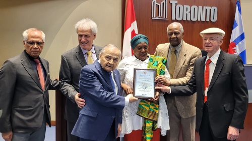 Group of six adults at an award ceremony in Toronto; a woman in colorful attire holds a certificate while others congratulate her.