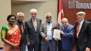 Six adults in formal attire pose in a conference room; a framed certificate is held at center, with a Canadian flag nearby.