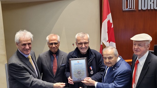 Five elderly men in suits pose for an award presentation, with a framed certificate held by the man in the center and a Turkish flag in the background.