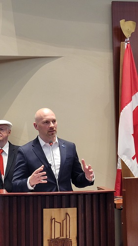 Bald man in a dark suit speaks at a wooden podium with a microphone, gesturing with his hands; an older man in a hat stands behind him, with a red-and-white flag in the background.