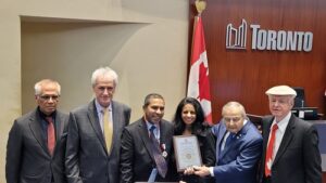 Six formally dressed individuals pose together in a Toronto office; a woman in the center holds a framed certificate, with a Canadian flag and a 'Toronto' sign behind them.