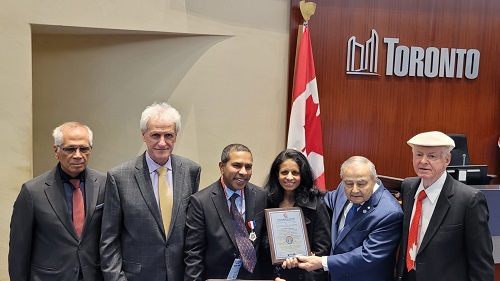Six formally dressed individuals pose together in a Toronto office; a woman in the center holds a framed certificate, with a Canadian flag and a 'Toronto' sign behind them.