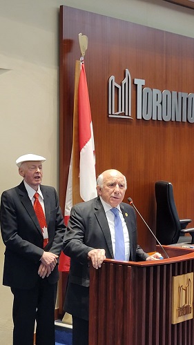 Two men in suits at a podium in a Toronto council chamber; one speaks into a microphone while the other stands nearby, with a Canadian flag and Toronto emblem visible in the background.