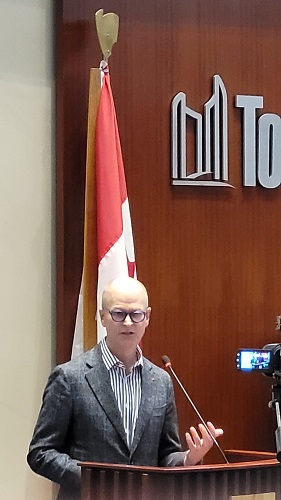 Bald man wearing glasses and a gray suit speaks at a podium, with a Canadian flag behind him in a conference room.