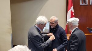Three men in a formal room as one pins a medal onto another during an award ceremony, with a flag in the background and a third man watching.