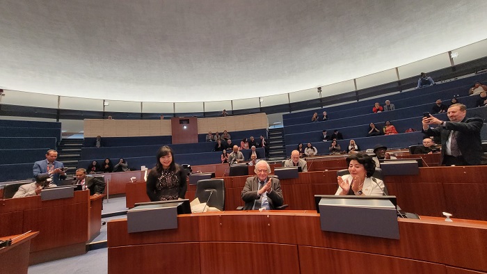 Legislative chamber with curved wooden desks and blue seating; a small group at the front applauds as spectators watch from the galleries.