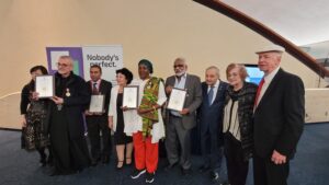 Group of people at an awards ceremony posing with certificates; backdrop features a banner that says "Nobody's perfect".