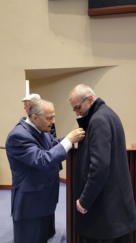 Two men in a room; one in a blue suit pins a badge onto the lapel of the man wearing a dark overcoat.