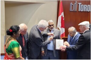 Group of six adults in formal attire at a ceremony; a man is handed a certificate as others look on, with a Canadian flag in the background.