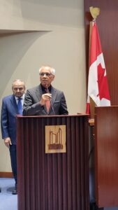 A man speaks at a wooden podium with a microphone while another man stands behind him; a Canadian flag is beside them in a formal indoor setting.