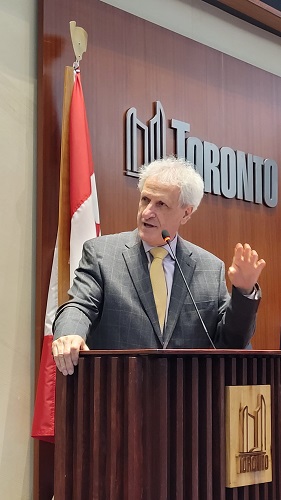 Older man in a gray suit and yellow tie giving a speech at a wooden podium, with a Canadian flag and a Toronto sign behind him.