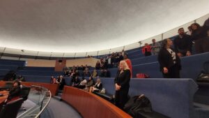 A woman in black standing at the front of a curved, blue-seat auditorium with attendees seated on stepped rows inside a modern lecture hall.
