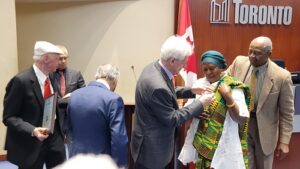 A formal ceremony at Toronto council; a man pins a medal on a woman wearing a green‑yellow traditional dress while others watch, with a Canadian flag in the background.