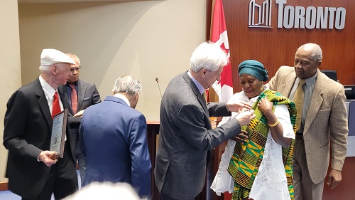 Dignitaries in a formal ceremony at a Toronto meeting room; a man adjusts a colorful shawl on an elderly woman while others watch.