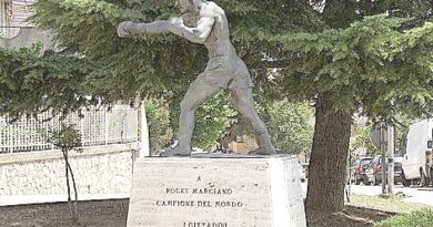 Statue of a muscular boxer in a punching stance on a pedestal with an inscription, in a tree-lined urban area.