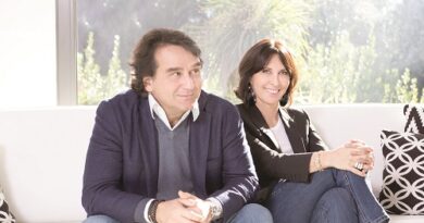 Two adults sit on a white couch in a bright room, smiling; man in a navy blazer and sweater, woman in a black jacket and jeans.