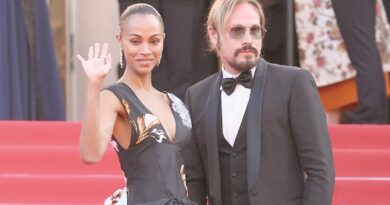 Woman in a black floral dress waves at the camera while standing next to a man in a black tuxedo on the red carpet.