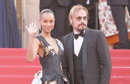 Woman in a black floral dress waves at the camera while standing next to a man in a black tuxedo on the red carpet.