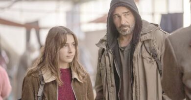 Two people stand side by side in a dim warehouse-like setting: a young woman with brown hair in a red sweater and a bearded man in a hooded jacket, looking ahead thoughtfully.