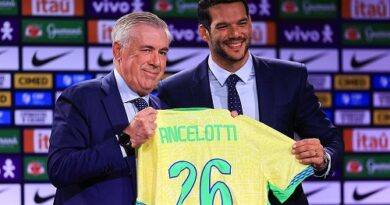 Two men in suits hold up a yellow soccer jersey that reads 'Ancelotti' with the number 26 at a press event.