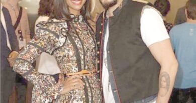 Smiling man and woman pose together at a social event; woman in a patterned dress, man in a white shirt with a dark vest, with other guests in the background.