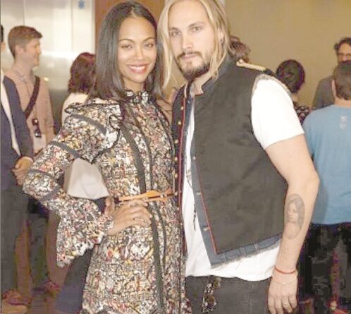 Smiling man and woman pose together at a social event; woman in a patterned dress, man in a white shirt with a dark vest, with other guests in the background.