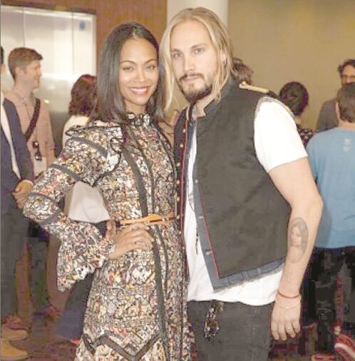 Smiling man and woman pose together at a social event; woman in a patterned dress, man in a white shirt with a dark vest, with other guests in the background.