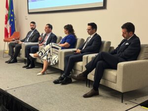 Panel of five speakers on stage in a conference room, with flags to the left and a projector screen above.