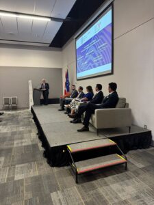 Panel discussion on a raised stage: presenter at a podium addressing five panelists seated on a sofa, with a projected slide on screen behind them.
