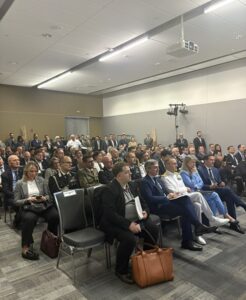 Audience seated in a conference room, many people in formal attire listening to a presentation, with a projector overhead and standing attendees in the back.