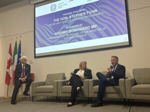 Three panelists sit on stage in a conference room; flags line the left wall and a presentation screen shows event details about Stephen Fuhr and Sherry Romano MP.