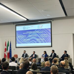 Panel discussion in a conference room with five speakers on stage, audience watching, and a projection screen showing the event title and logos; flags on the left.