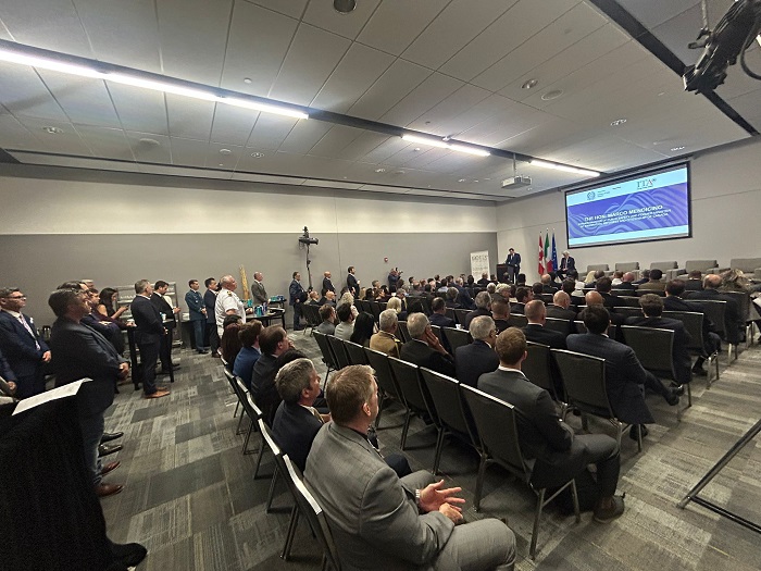 Large conference room with a speaker at a podium addressing a seated audience; presentation screen and flags in the background