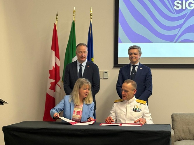 Woman in a light blue blazer signs documents at a table beside a man in a white naval uniform; two suited men stand behind, with Canadian and other flags in the background and a presentation screen to the right.