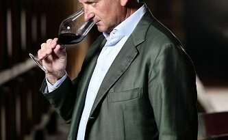 A middle-aged man in a green suit tastes red wine from a stemmed glass in a dim wine cellar with barrels lined up along the wall.