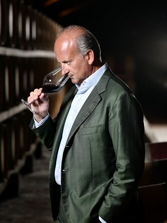 A middle-aged man in a green suit tastes red wine from a stemmed glass in a dim wine cellar with barrels lined up along the wall.
