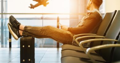 Man sitting in an airport departure lounge with a backpacking suitcase, legs stretched out, watching a plane take off through the window.