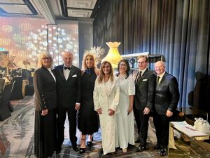 Seven adults in formal attire pose together at a black-tie gala, with gold decor and a large lamp in the background.