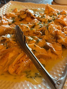 Shredded chicken in orange sauce with herbs on a white plate, a metal tong resting beside it.