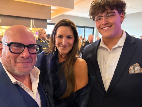 Three people posing at a formal event: a bald man with glasses, a woman with long brown hair, and a young man in a suit smiling nearby.
