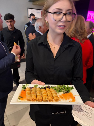 A woman in a black shirt holds a platter of golden spring rolls with orange dipping sauce at a social event.