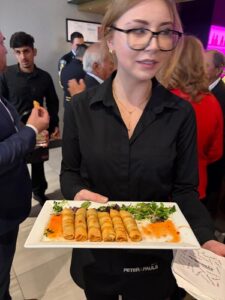 Waitress in a black shirt presents a plate of six spring rolls with orange sauce and greens at a social event.