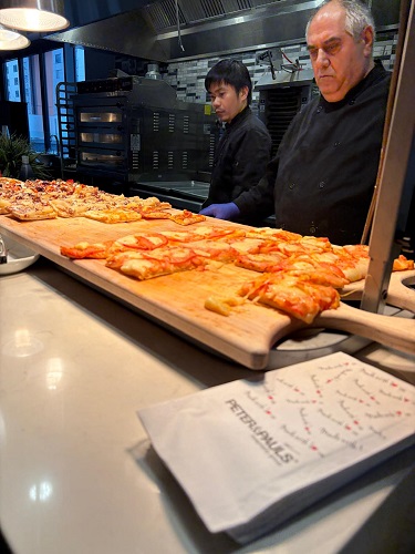 Two chefs work behind the counter as freshly baked pizzas rest on wooden boards in a busy kitchen.