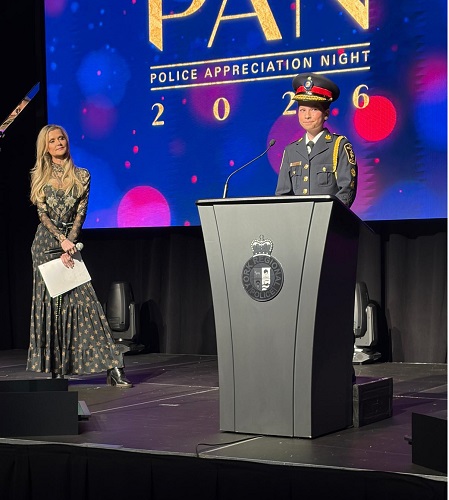 Police officer in formal uniform at a podium giving a speech during Police Appreciation Night, with a female presenter holding notes beside the stage.