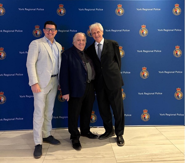 Three men pose together in front of a blue York Regional Police backdrop; left man wears a light gray suit, middle man a dark blazer and black shirt, right man a black suit and tie, all smiling.
