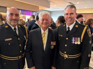 Three men posing together at a formal event; two in dress uniforms with medals flank an older man in a suit and yellow tie, all smiling.